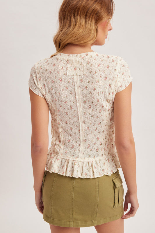 A short-sleeve light pink ditsy floral tee featuring a fully smocked body, a round neckline, and lettuce-edge trim at the sleeves and hem.