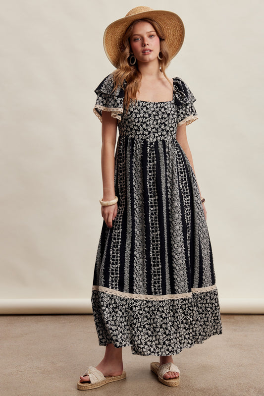 A black floral border print maxi dress featuring a square neckline, smocked bodice, tiered flutter sleeves with lace trim, and functional side pockets.