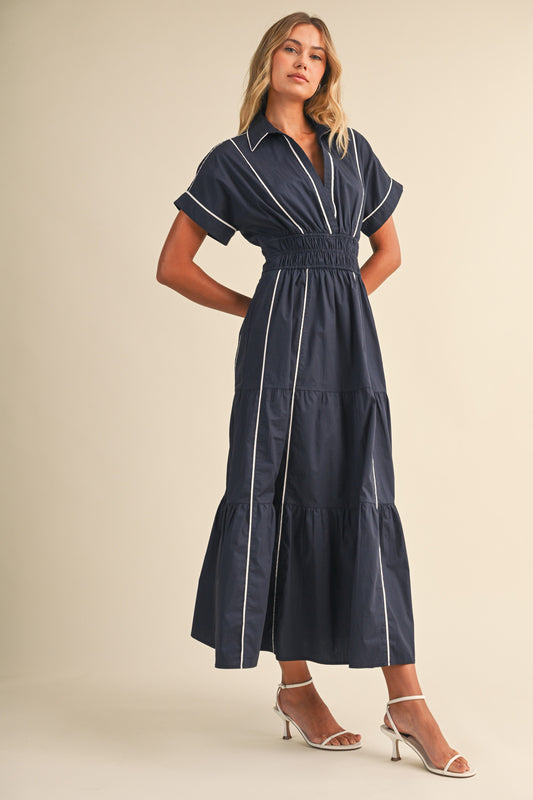 A short-sleeve navy blue shirt maxi dress featuring a classic collar, white vertical contrast piping, a thick smocked elastic waistband, and a tiered A-line skirt.