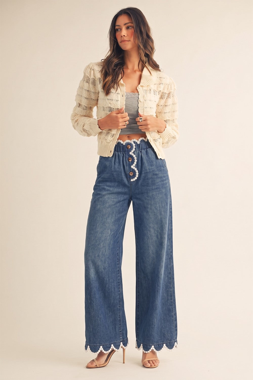 A pair of women's high-rise draped wide-leg denim pants in a deep indigo wash featuring a gathered elastic paperbag waistband, a button-front fly, functional side pockets, and a white daisy-edge scallop lace trim along the waist and bottom hems.
