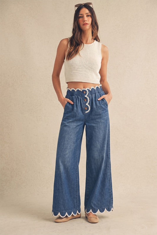 A pair of women's high-rise draped wide-leg denim pants in a deep indigo wash featuring a gathered elastic paperbag waistband, a button-front fly, functional side pockets, and a white daisy-edge scallop lace trim along the waist and bottom hems.