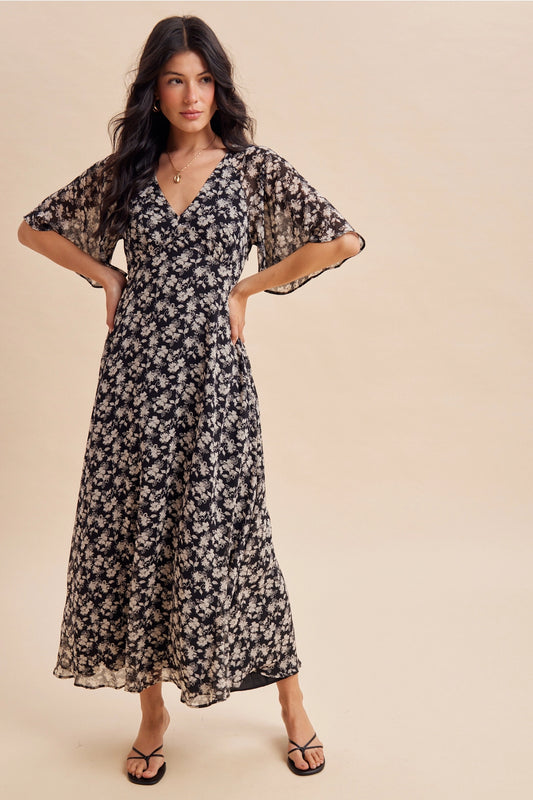 A women's v-neck maxi dress in a black and cream floral print featuring short flutter sleeves and an empire waist silhouette.
