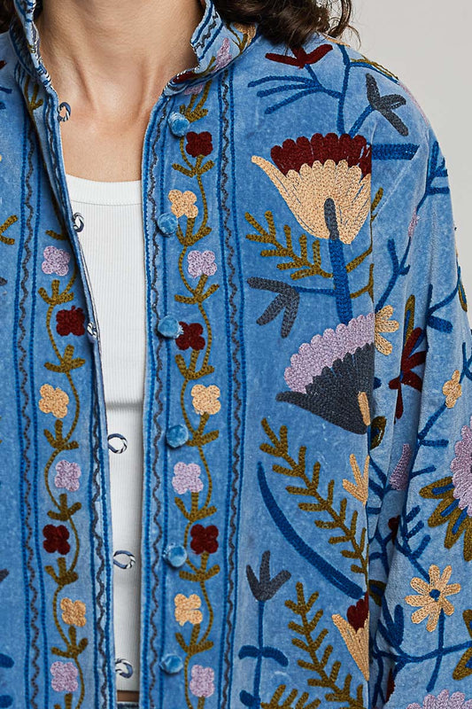 A women's dusty denim blue long-sleeve velvet jacket with allover folk floral embroidery in rust, yellow, and lavender, styled with a mandarin collar, wide cuffs, and front loop buttons.
