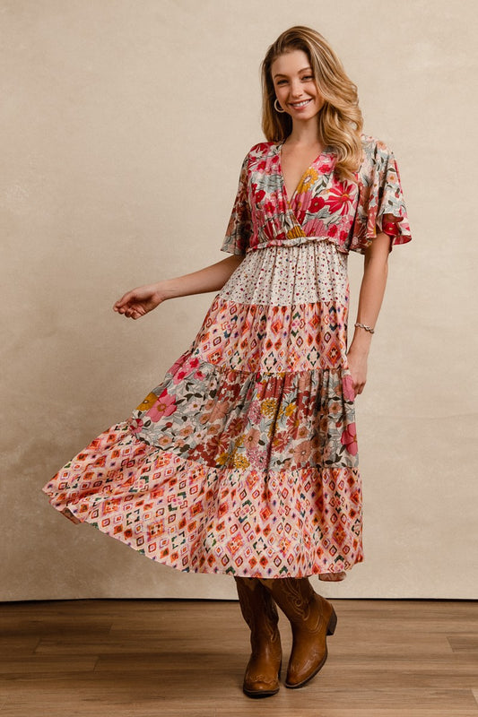 A short-sleeve tiered patchwork midi dress in a rose pink floral and geometric print featuring a deep V-neckline, flutter sleeves, and an elastic waist.