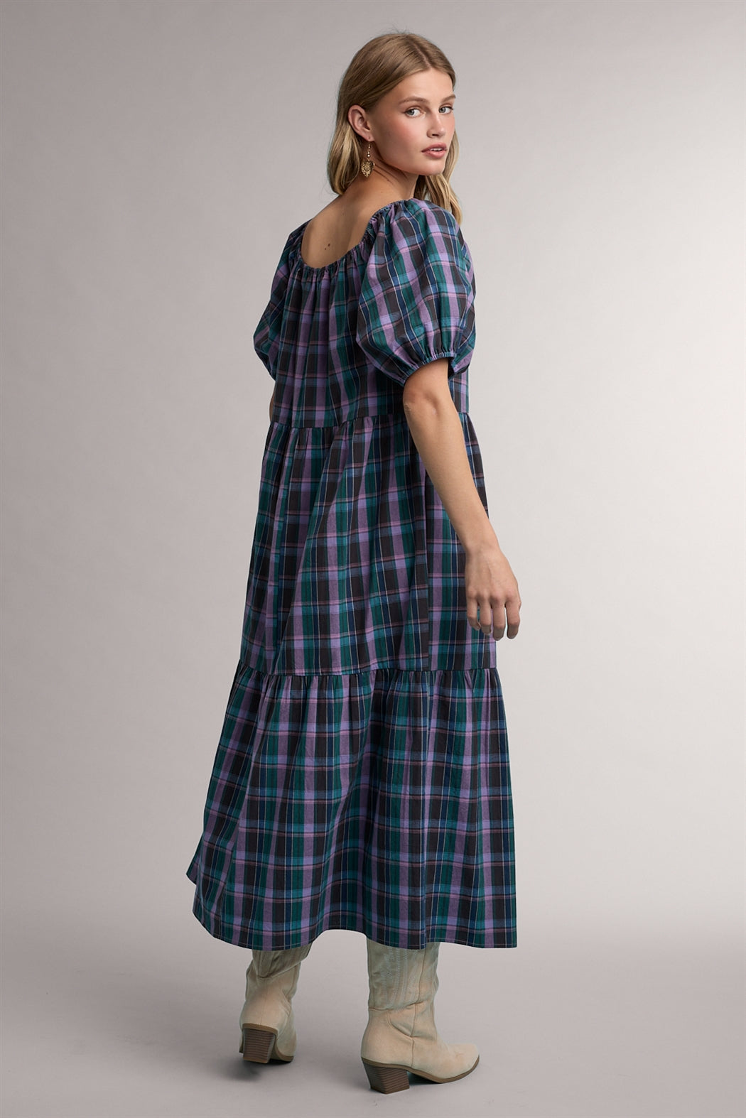 A women's tiered midi-length dress in a green and violet plaid pattern featuring short puff sleeves, a gathered scoop neck with a tie detail, and functional side pockets.