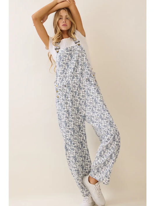 A women's sleeveless floral printed cropped overall jumpsuit in a blue and cream pattern featuring adjustable buckle shoulder straps, side button closures, utility slash pockets, and a relaxed wide-leg silhouette worn over a white short-sleeve tee.