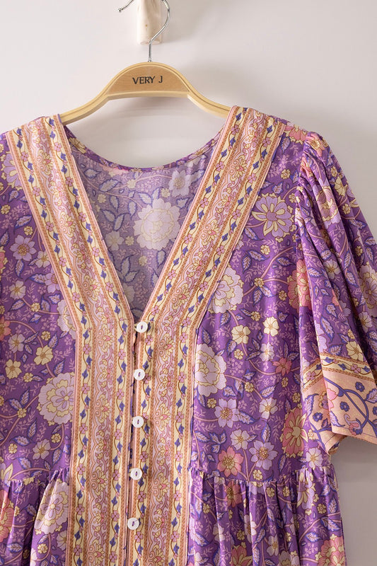 A short-sleeve tiered floral maxi dress in a lavender, coral, and gold mix featuring a deep V-neckline, button-down front closure, and decorative border print detailing.