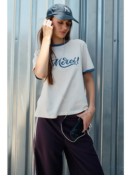 A women's light sand crewneck ringer t-shirt featuring deep blue contrast trim along the neckline and sleeves, short sleeves, and cursive blue embroidered script across the chest that says, Merci.