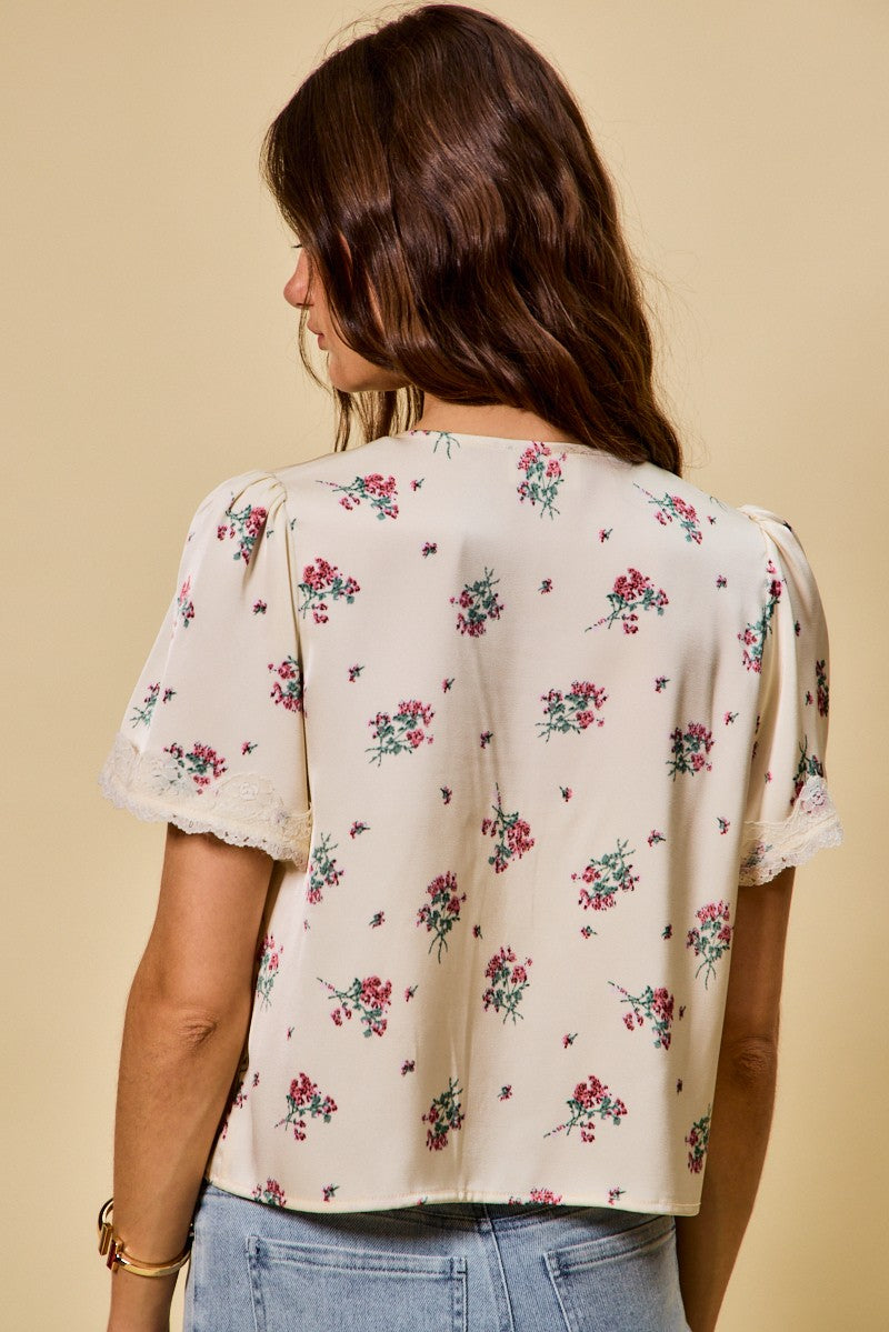 A short-sleeve ivory satin floral print blouse featuring a functional double tie-front closure, puff sleeves, and scalloped lace trim along the neckline and hem.