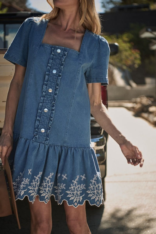 A women's medium wash blue denim short-sleeve mini dress featuring a square neckline, ruffled button placket, and cream floral embroidered scalloped hemline.