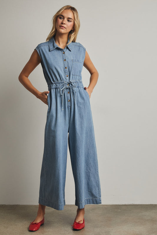 A cap-sleeve light wash denim jumpsuit featuring a classic button-down front, an adjustable drawstring waist, and a relaxed wide-leg fit.