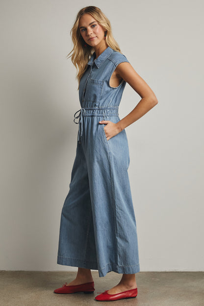 A cap-sleeve light wash denim jumpsuit featuring a classic button-down front, an adjustable drawstring waist, and a relaxed wide-leg fit.