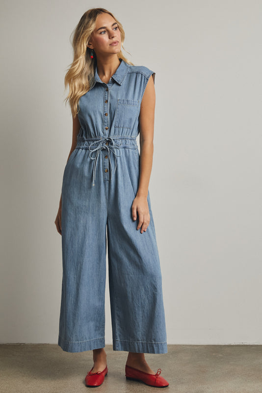 A cap-sleeve light wash denim jumpsuit featuring a classic button-down front, an adjustable drawstring waist, and a relaxed wide-leg fit.