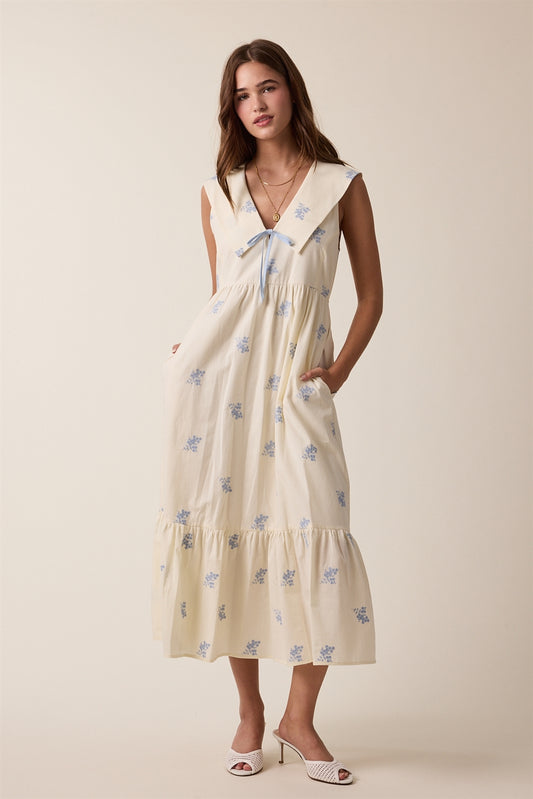 Cream sleeveless midi dress with floral design. A slight hooded peter-pan collar and a front blue accent tie. Featuring side pockets. 