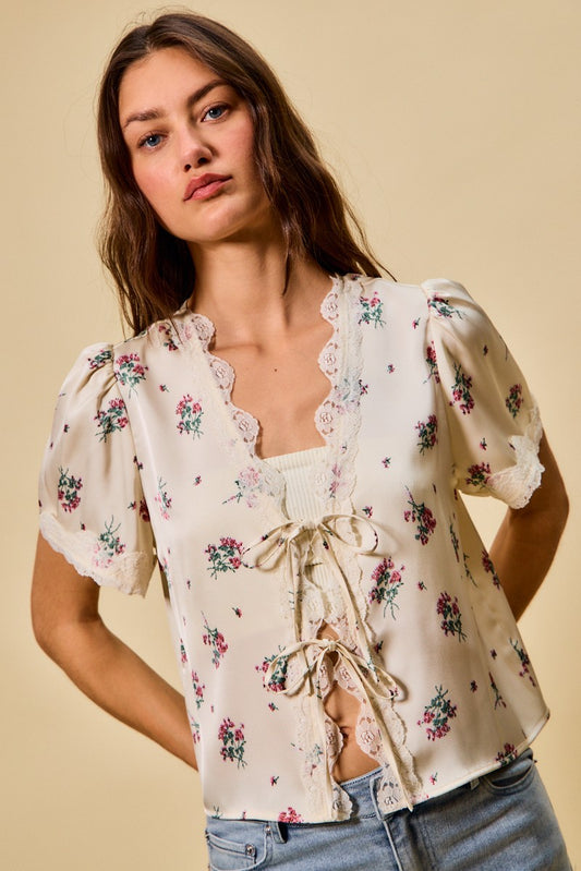 A short-sleeve ivory satin floral print blouse featuring a functional double tie-front closure, puff sleeves, and scalloped lace trim along the neckline and hem.