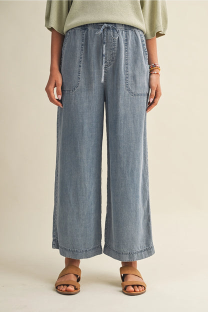 A pair of women's high-waisted wide-leg denim chambray pants featuring a gathered elastic drawstring waistband and functional front patch pockets.
