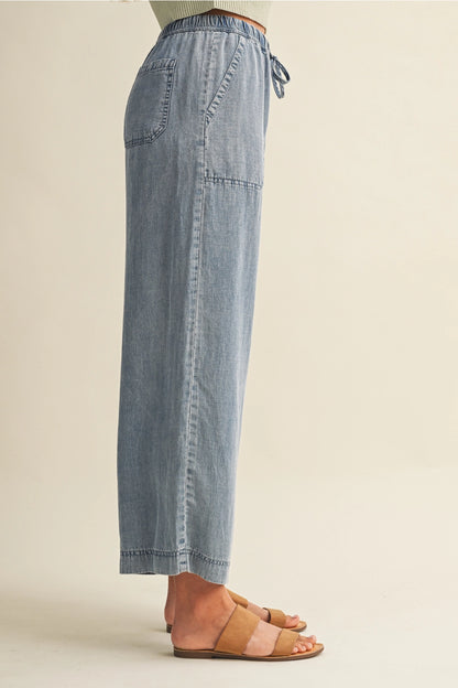 A pair of women's high-waisted wide-leg denim chambray pants featuring a gathered elastic drawstring waistband and functional front patch pockets.