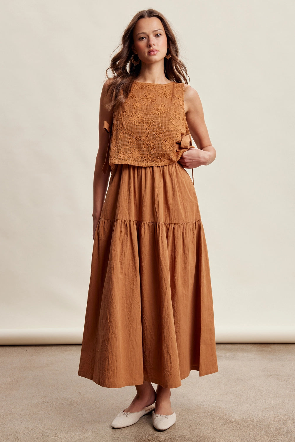A women's brown sleeveless tiered maxi dress featuring a tonal embroidered floral lace vest overlay with adjustable side tie details.
