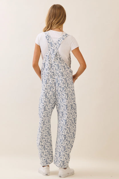 A women's sleeveless floral printed cropped overall jumpsuit in a blue and cream pattern featuring adjustable buckle shoulder straps, side button closures, utility slash pockets, and a relaxed wide-leg silhouette worn over a white short-sleeve tee.