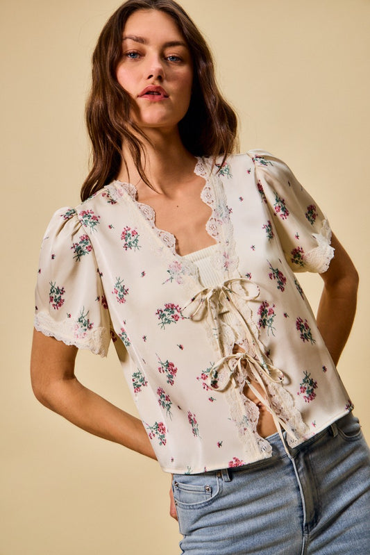 A short-sleeve ivory satin floral print blouse featuring a functional double tie-front closure, puff sleeves, and scalloped lace trim along the neckline and hem.