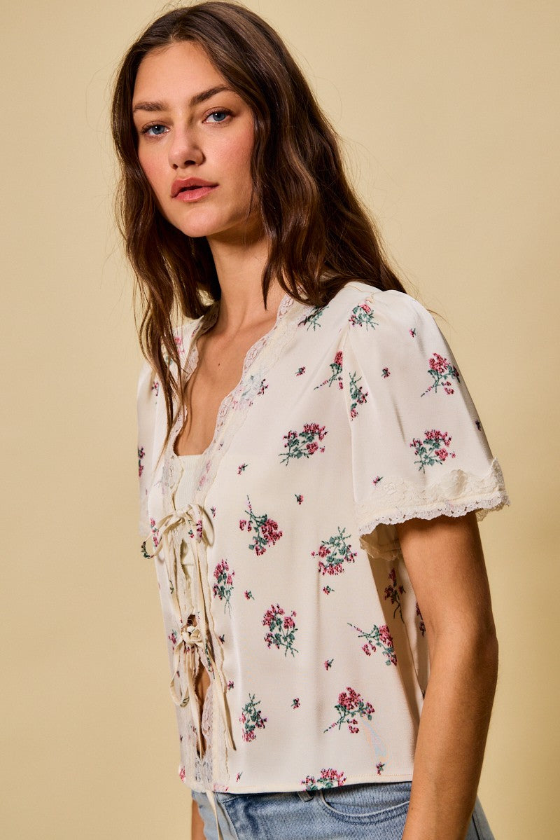 A short-sleeve ivory satin floral print blouse featuring a functional double tie-front closure, puff sleeves, and scalloped lace trim along the neckline and hem.