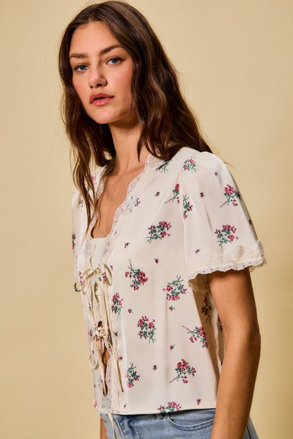 A short-sleeve ivory satin floral print blouse featuring a functional double tie-front closure, puff sleeves, and scalloped lace trim along the neckline and hem.