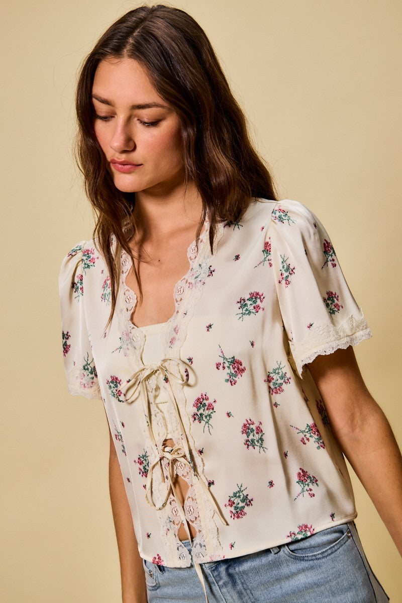 A short-sleeve ivory satin floral print blouse featuring a functional double tie-front closure, puff sleeves, and scalloped lace trim along the neckline and hem.