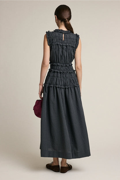A women's sleeveless maxi dress in a dark gray and black plaid pattern featuring a high ruffled neckline, a fully elasticized smocked bodice, and a flowing tiered skirt with delicate ruffle trim.