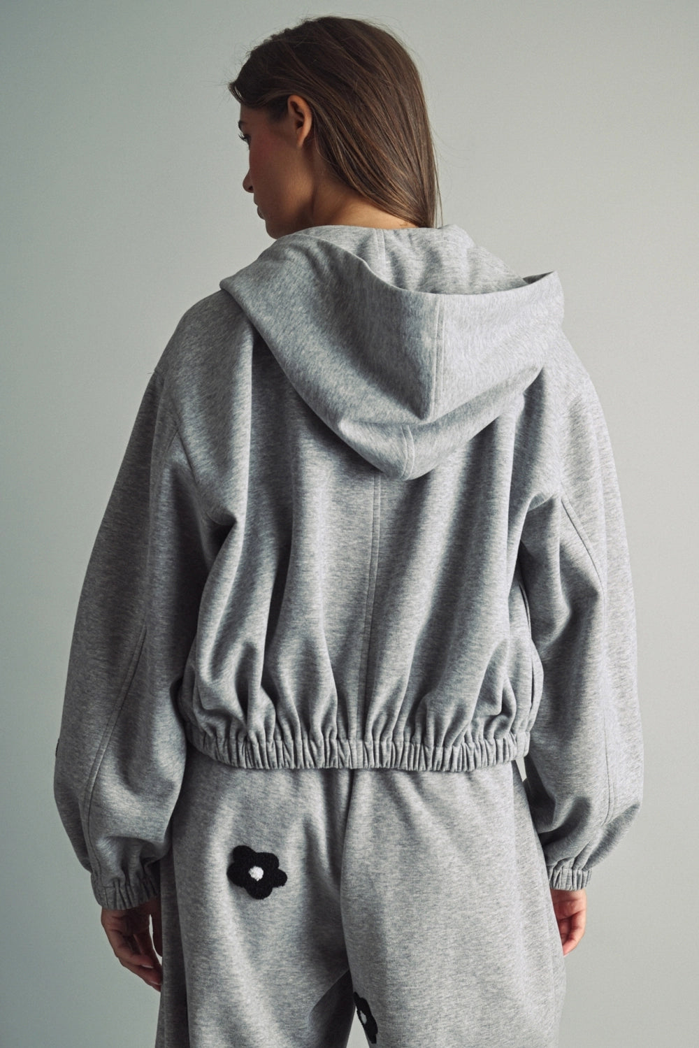 A women's heather gray full-zip hooded fleece jacket featuring dropped shoulders, ribbed hem cuffs, and black chenille flower patches on the front and sleeves.