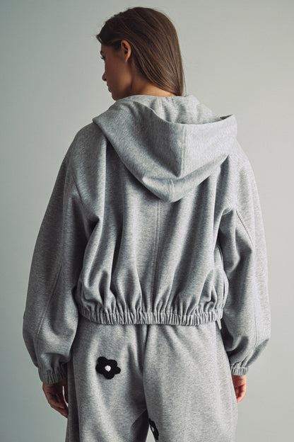 A women's heather gray full-zip hooded fleece jacket featuring dropped shoulders, ribbed hem cuffs, and black chenille flower patches on the front and sleeves.