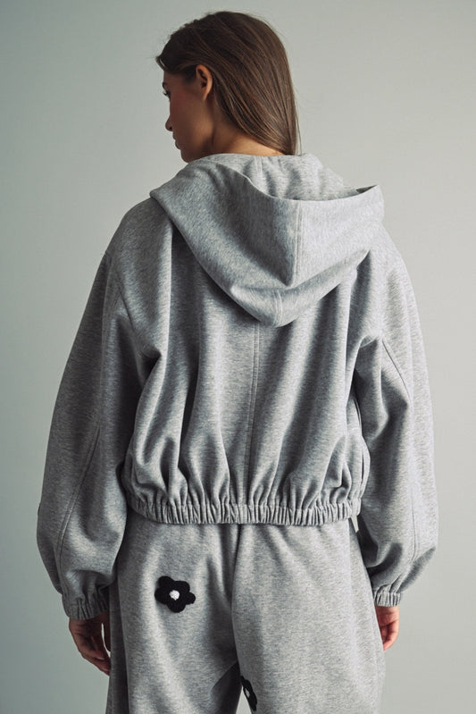 A women's heather gray full-zip hooded fleece jacket featuring dropped shoulders, ribbed hem cuffs, and black chenille flower patches on the front and sleeves.