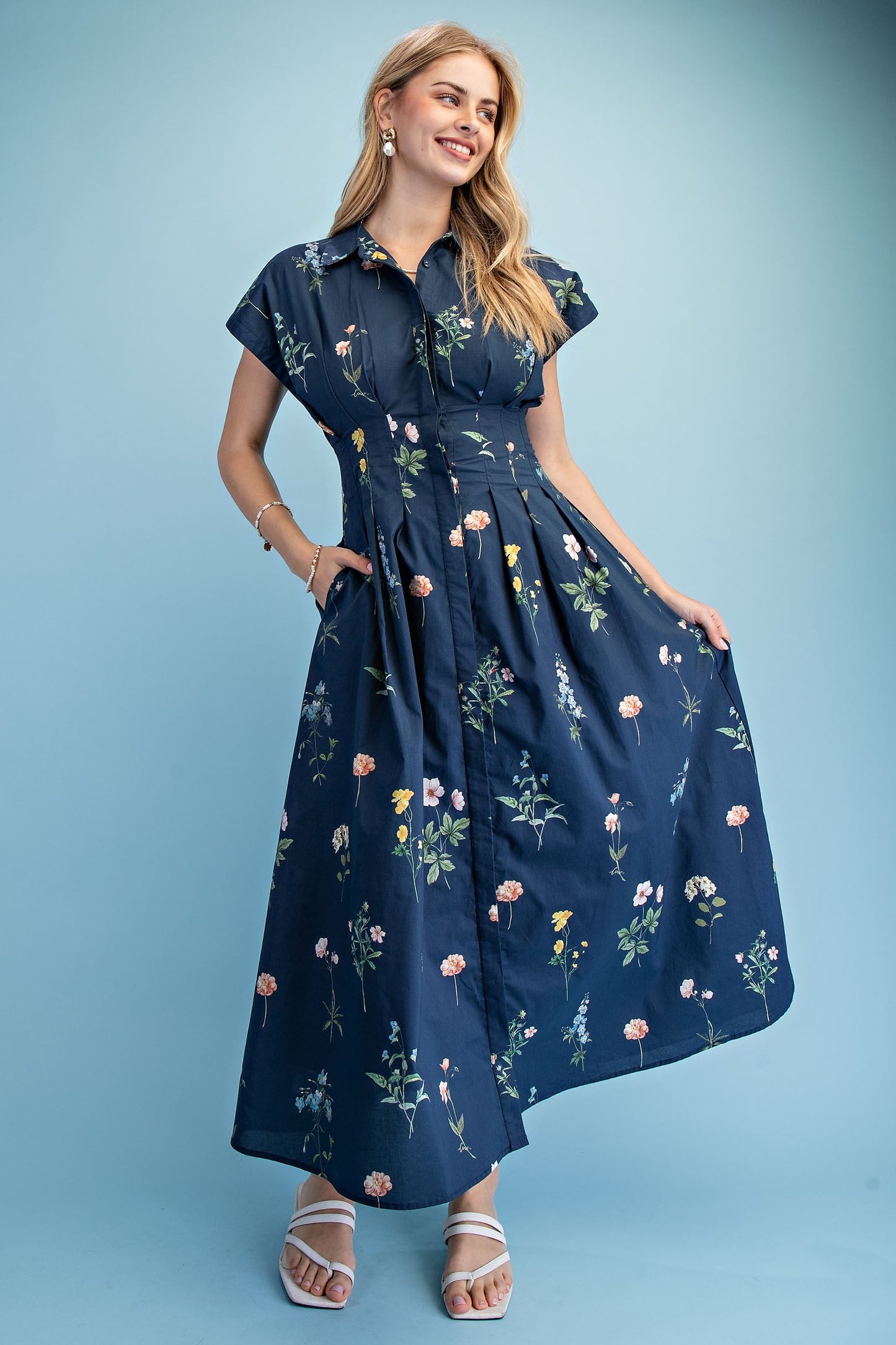 A short-sleeve button-up maxi dress in a navy blue floral wildflower print featuring a classic collar, deep side pockets, and detailed pleating around the waistline.