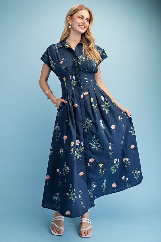 A short-sleeve button-up maxi dress in a navy blue floral wildflower print featuring a classic collar, deep side pockets, and detailed pleating around the waistline.