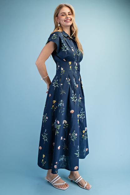 A short-sleeve button-up maxi dress in a navy blue floral wildflower print featuring a classic collar, deep side pockets, and detailed pleating around the waistline.