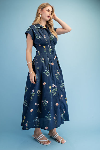 A short-sleeve button-up maxi dress in a navy blue floral wildflower print featuring a classic collar, deep side pockets, and detailed pleating around the waistline.