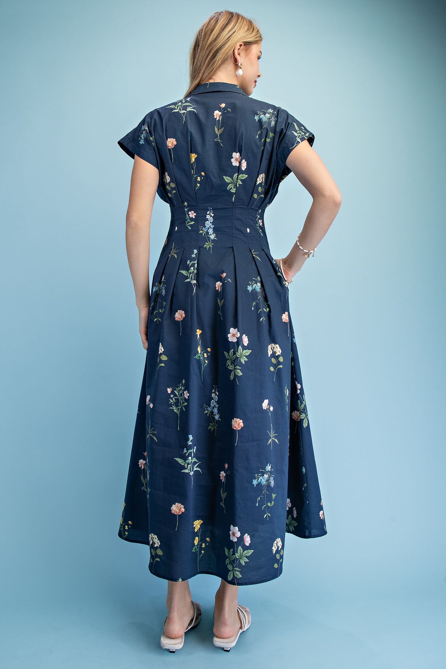 A short-sleeve button-up maxi dress in a navy blue floral wildflower print featuring a classic collar, deep side pockets, and detailed pleating around the waistline.