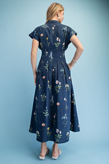 A short-sleeve button-up maxi dress in a navy blue floral wildflower print featuring a classic collar, deep side pockets, and detailed pleating around the waistline.