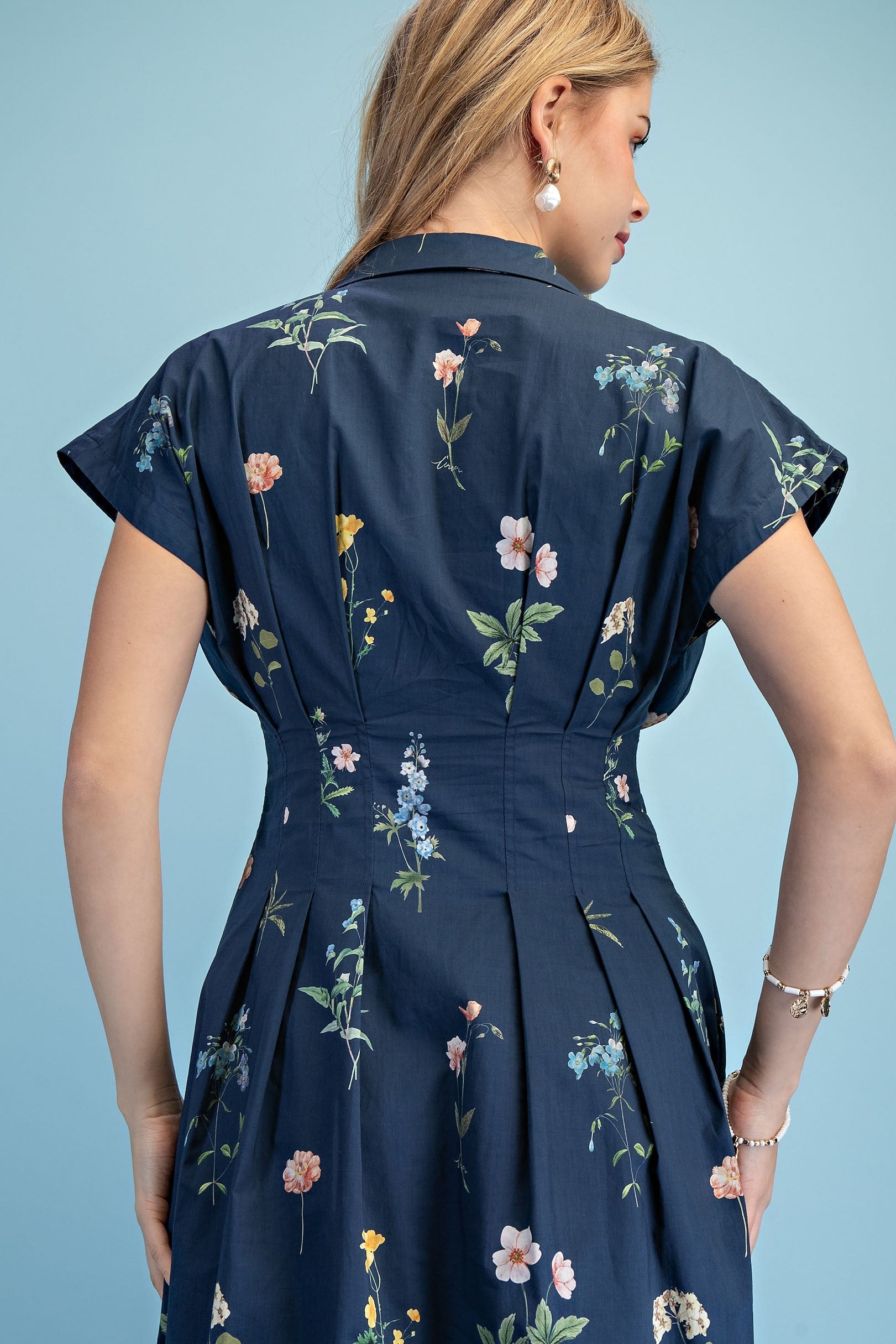 A short-sleeve button-up maxi dress in a navy blue floral wildflower print featuring a classic collar, deep side pockets, and detailed pleating around the waistline.