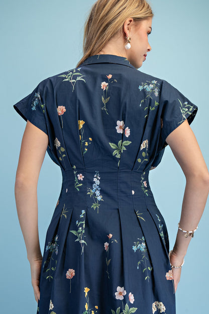 A short-sleeve button-up maxi dress in a navy blue floral wildflower print featuring a classic collar, deep side pockets, and detailed pleating around the waistline.