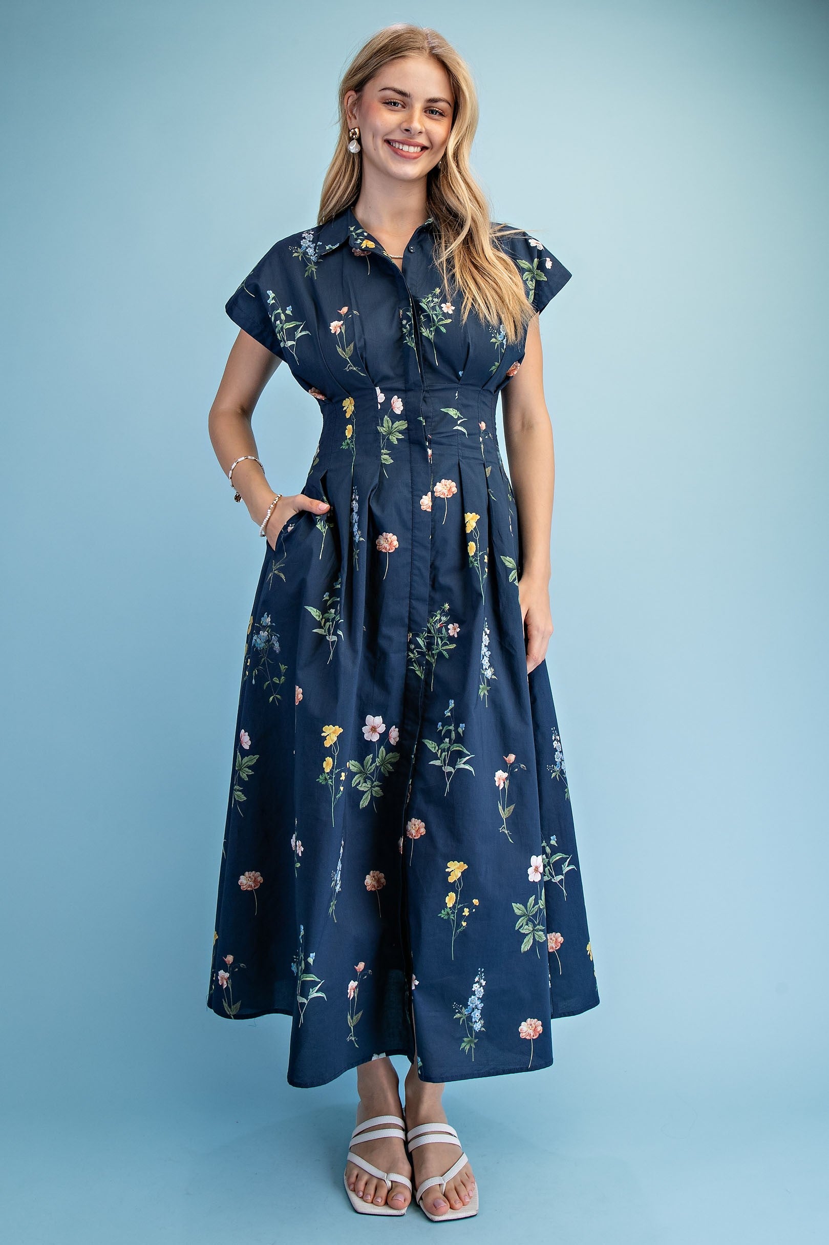 A short-sleeve button-up maxi dress in a navy blue floral wildflower print featuring a classic collar, deep side pockets, and detailed pleating around the waistline.