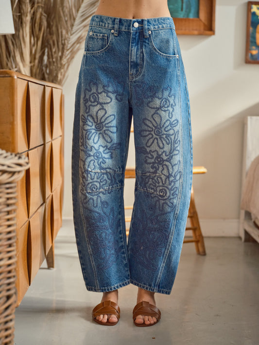A pair of high-waisted women's barrel jeans in a dark wash featuring intricate tonal blue floral embroidery down the front of the curved legs.
