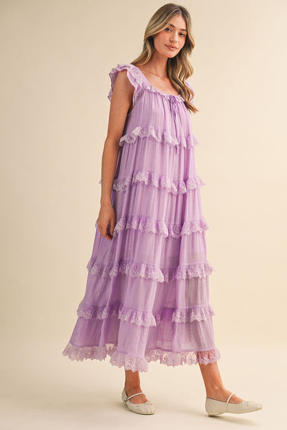 A sleeveless tiered maxi dress in lavender featuring intricate scalloped lace trim, a scooped neckline with an adjustable front tie, and flutter shoulder straps.