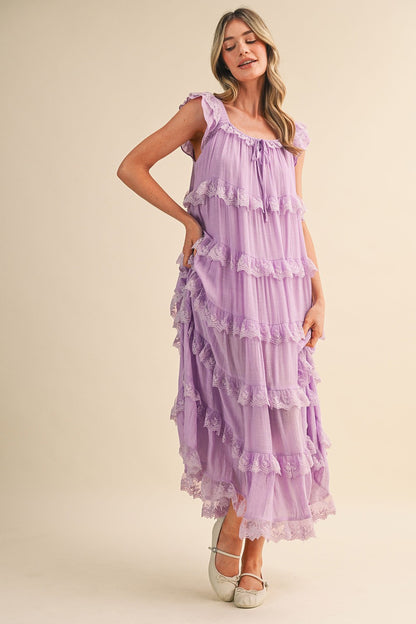 A sleeveless tiered maxi dress in lavender featuring intricate scalloped lace trim, a scooped neckline with an adjustable front tie, and flutter shoulder straps.
