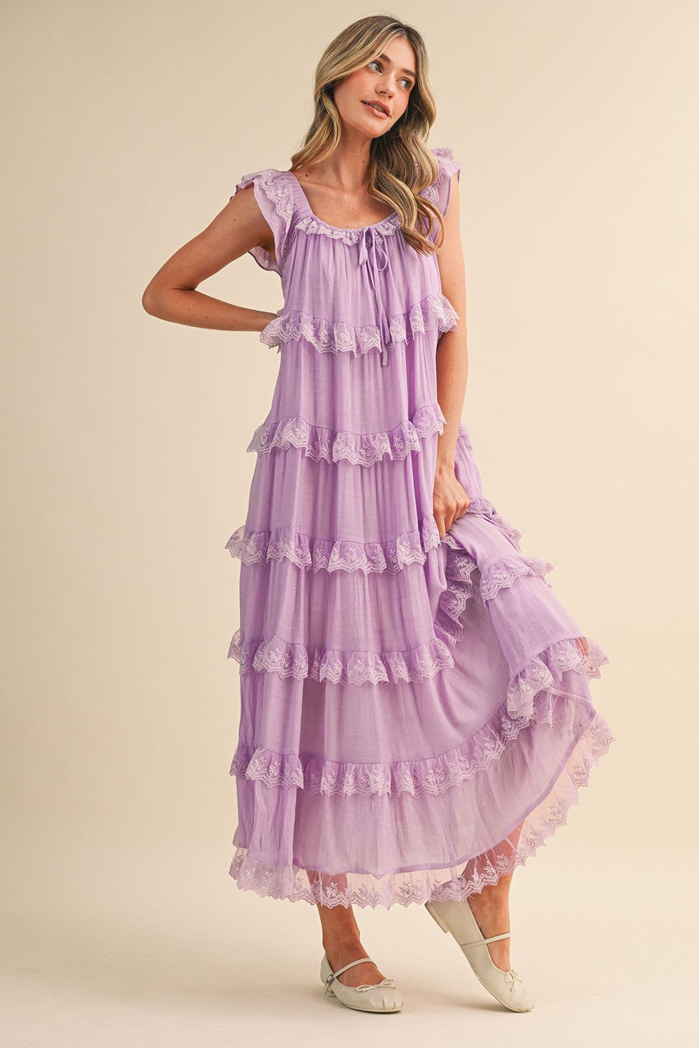 A sleeveless tiered maxi dress in lavender featuring intricate scalloped lace trim, a scooped neckline with an adjustable front tie, and flutter shoulder straps.