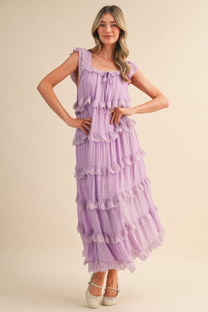 A sleeveless tiered maxi dress in lavender featuring intricate scalloped lace trim, a scooped neckline with an adjustable front tie, and flutter shoulder straps.