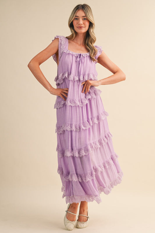 A sleeveless tiered maxi dress in lavender featuring intricate scalloped lace trim, a scooped neckline with an adjustable front tie, and flutter shoulder straps.