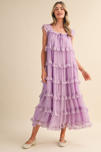 A sleeveless tiered maxi dress in lavender featuring intricate scalloped lace trim, a scooped neckline with an adjustable front tie, and flutter shoulder straps.