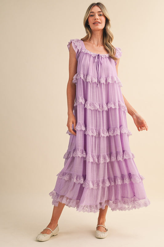 A sleeveless tiered maxi dress in lavender featuring intricate scalloped lace trim, a scooped neckline with an adjustable front tie, and flutter shoulder straps.