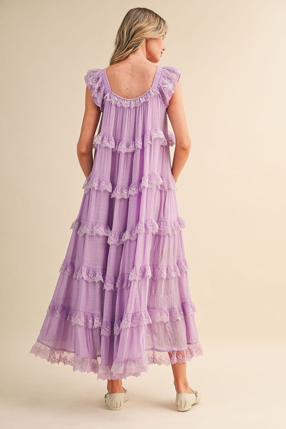 A sleeveless tiered maxi dress in lavender featuring intricate scalloped lace trim, a scooped neckline with an adjustable front tie, and flutter shoulder straps.