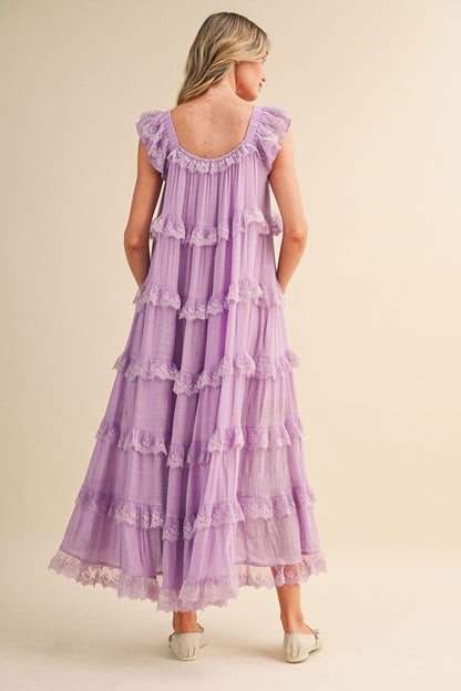 A sleeveless tiered maxi dress in lavender featuring intricate scalloped lace trim, a scooped neckline with an adjustable front tie, and flutter shoulder straps.